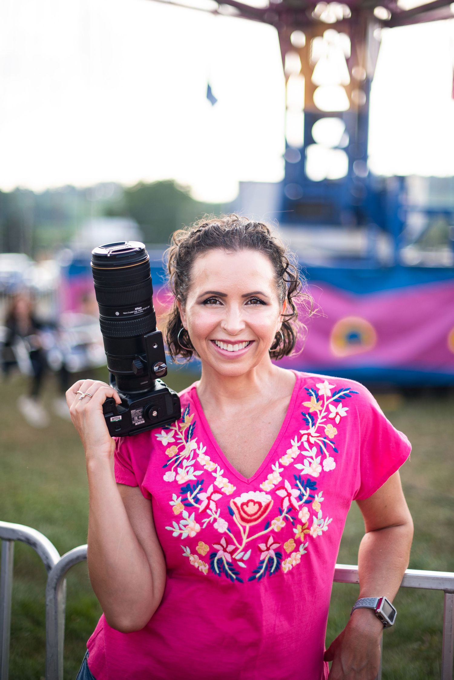 Photographer in pink embroidered shirt smiles while holding professional camera equipment at outdoor event.