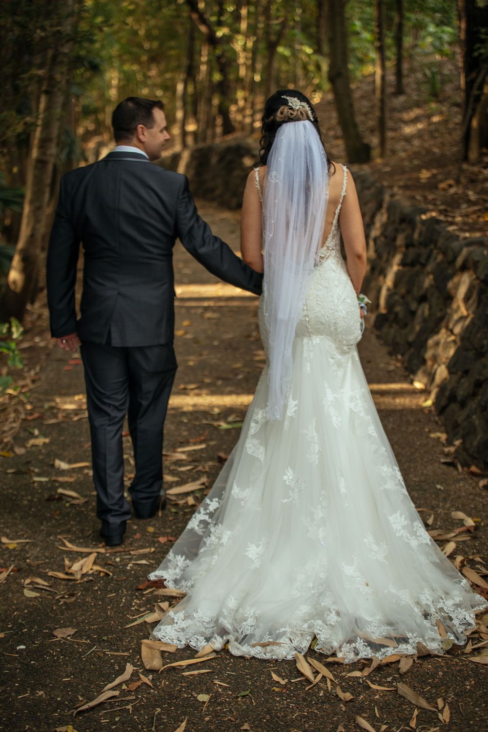 Wedding couple walks hand in hand down wooded forest path in elegant attire.
