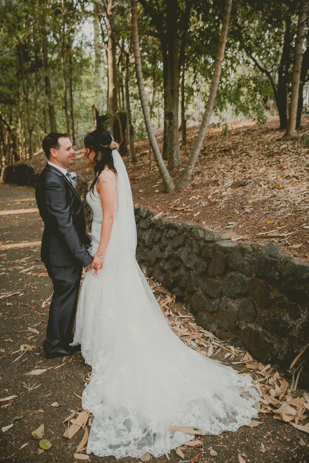 Elegant full-length wedding portrait beside rustic stone wall in woodland setting.
