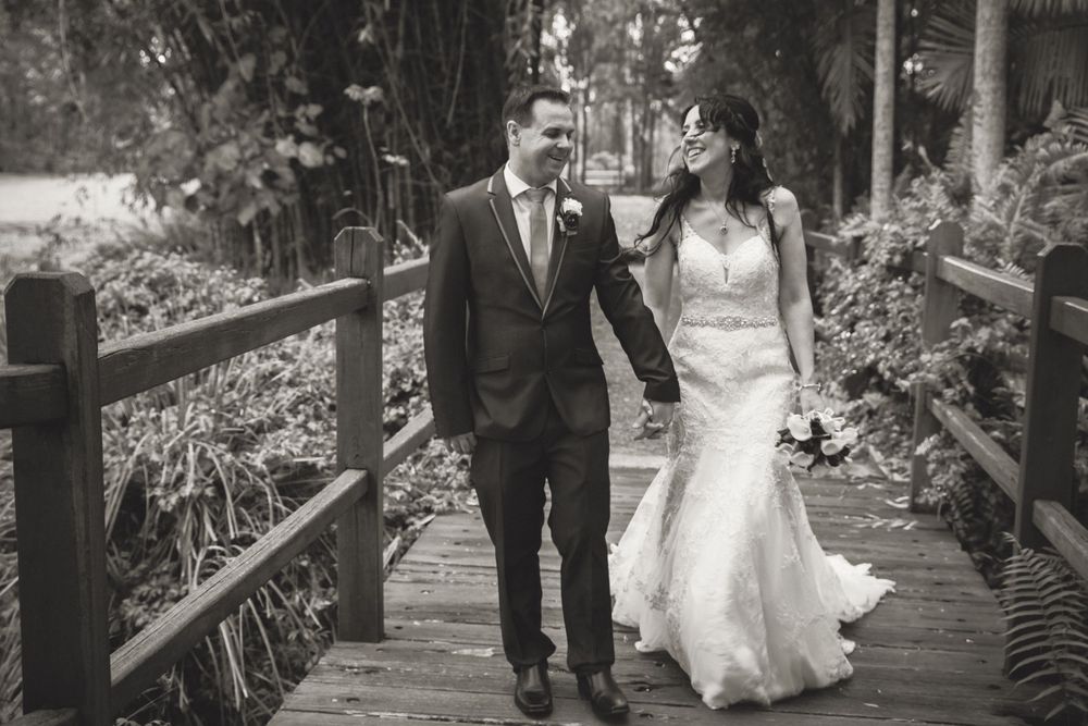 Black and white wedding photo of couple walking together on wooden bridge surrounded by nature.