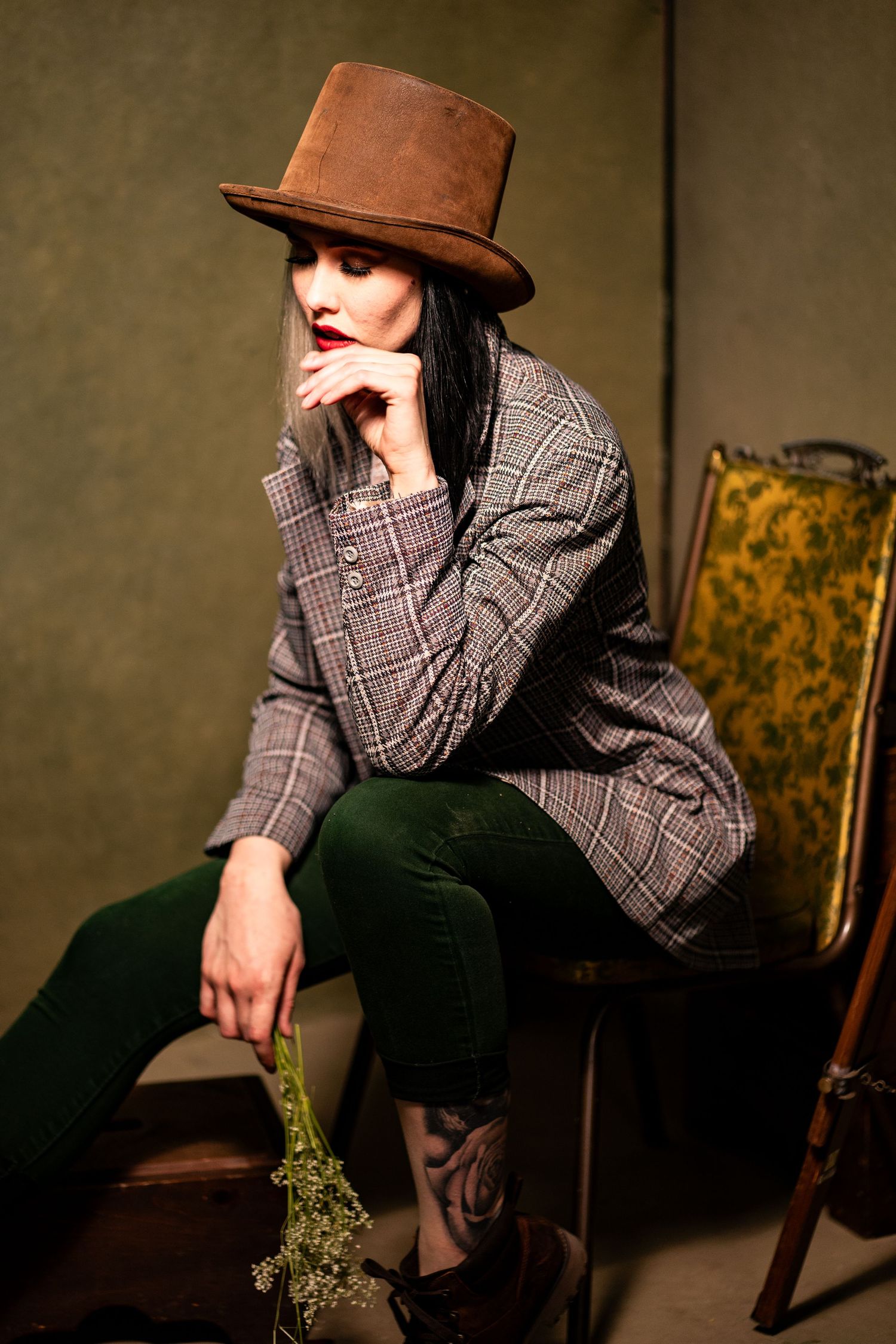 A stylish portrait of someone in a brown top hat and plaid blazer holding dried flowers.
