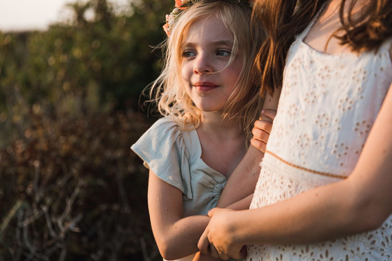 Young girl in blue dress hugging her mother outdoors in warm golden sunlight among green foliage.