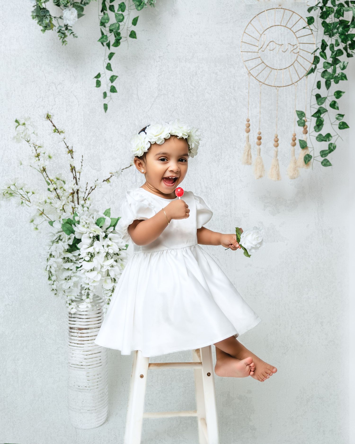 Joyful toddler in white dress and floral crown sitting on stool against decorative wall backdrop.