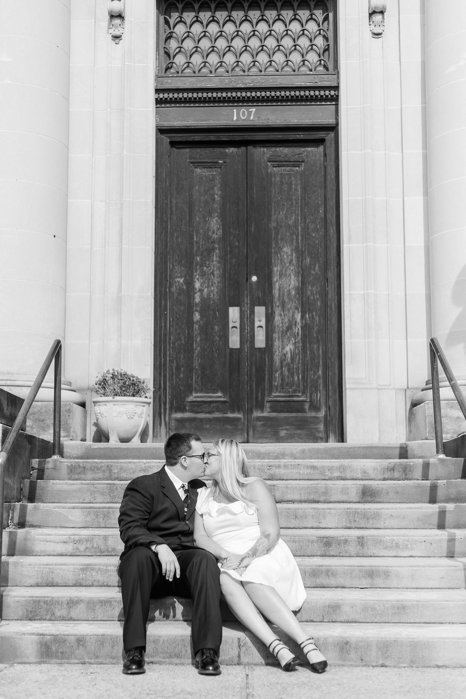 A romantic couple sits on stone steps in front of a vintage wooden door in formal attire.