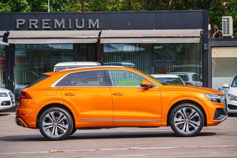 Side profile of an orange Audi Q8 SUV parked in front of a Premium dealership.