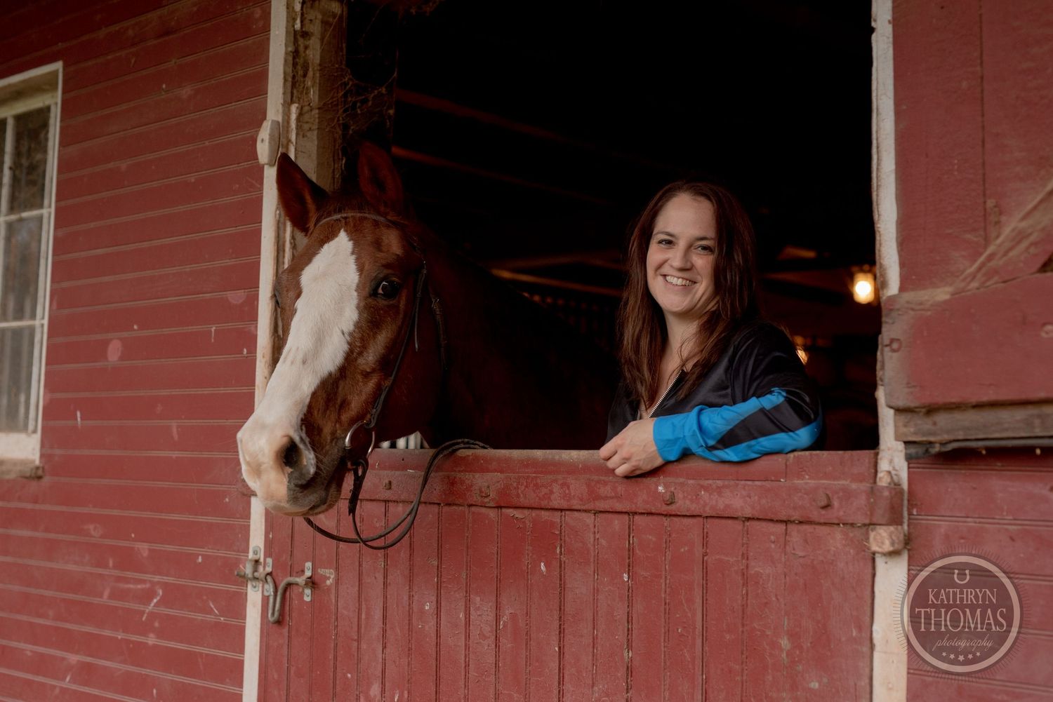 A horse with a white blaze looks out from a red barn window during an equestrian photoshoot.