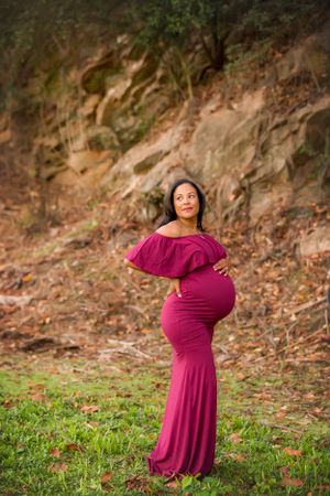 Pregnant woman in burgundy dress standing in green grass with rock formation behind her.