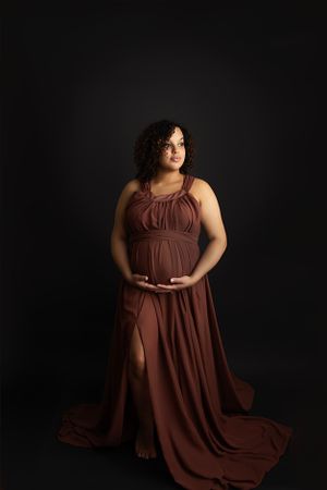Pregnant mom standing on a black backdrop wearing a burgundy maternity gown cradling her baby bump.