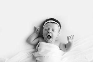 Newborn baby girl in a white wrap and flower headband yawning during her session