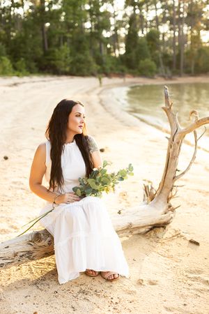 Woman sitting on beech wood wearing a white dress, holding flowers, looking towards Clarks Hill Lake.