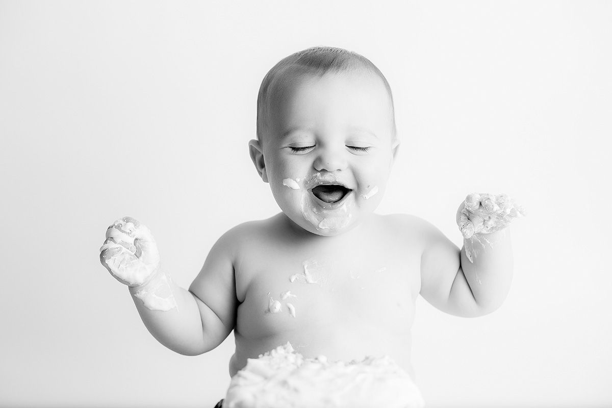 One year old baby boy enjoying birthday cake laughing with cake on his face.