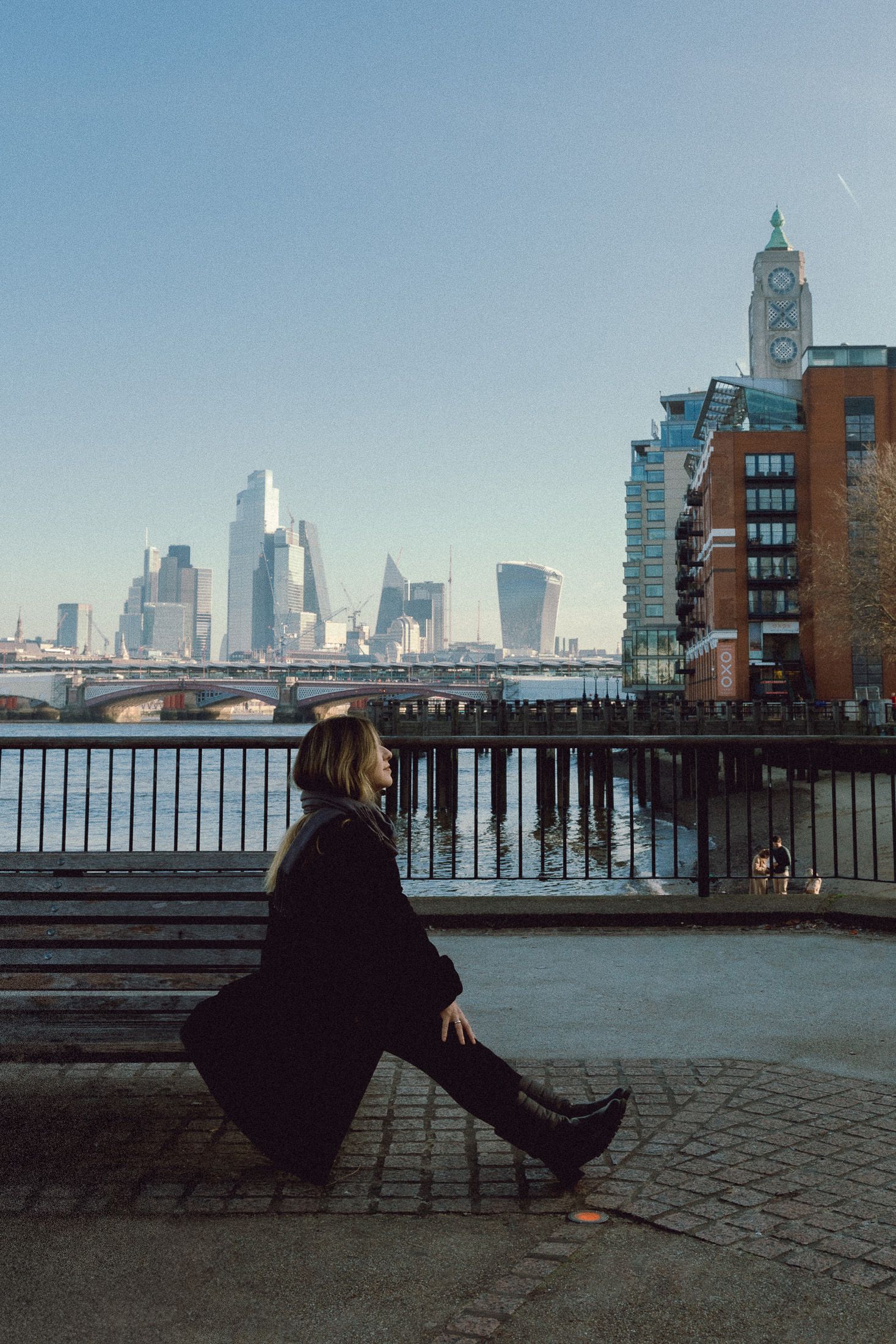 Silhouetted figure relaxes by Thames River with London skyscrapers and bridge visible in distance.