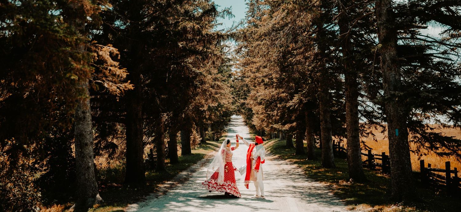 Couple in red traditional attire walking down tree-lined path.