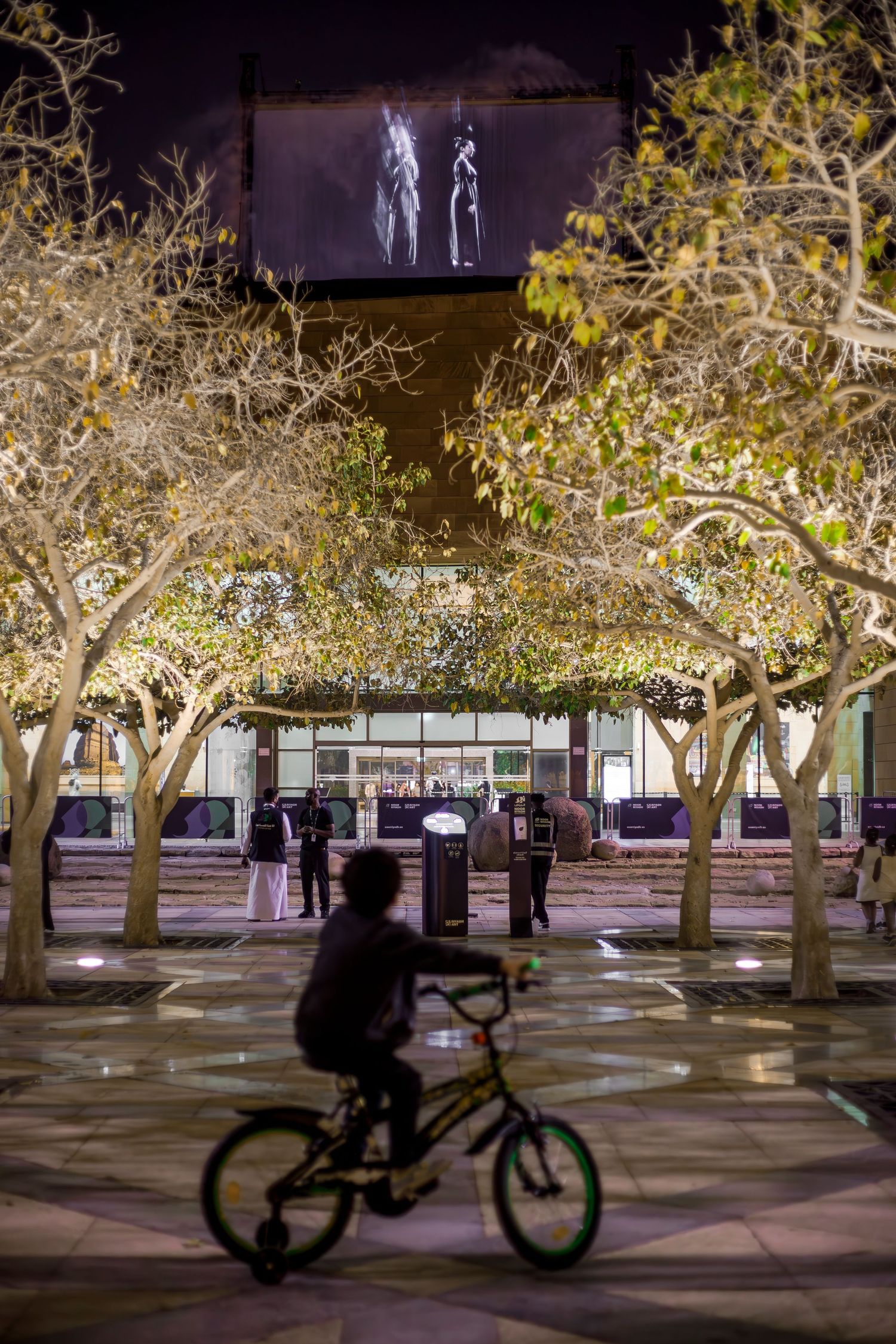 Black and white projection on screen above lit trees with silhouetted cyclist passing below.