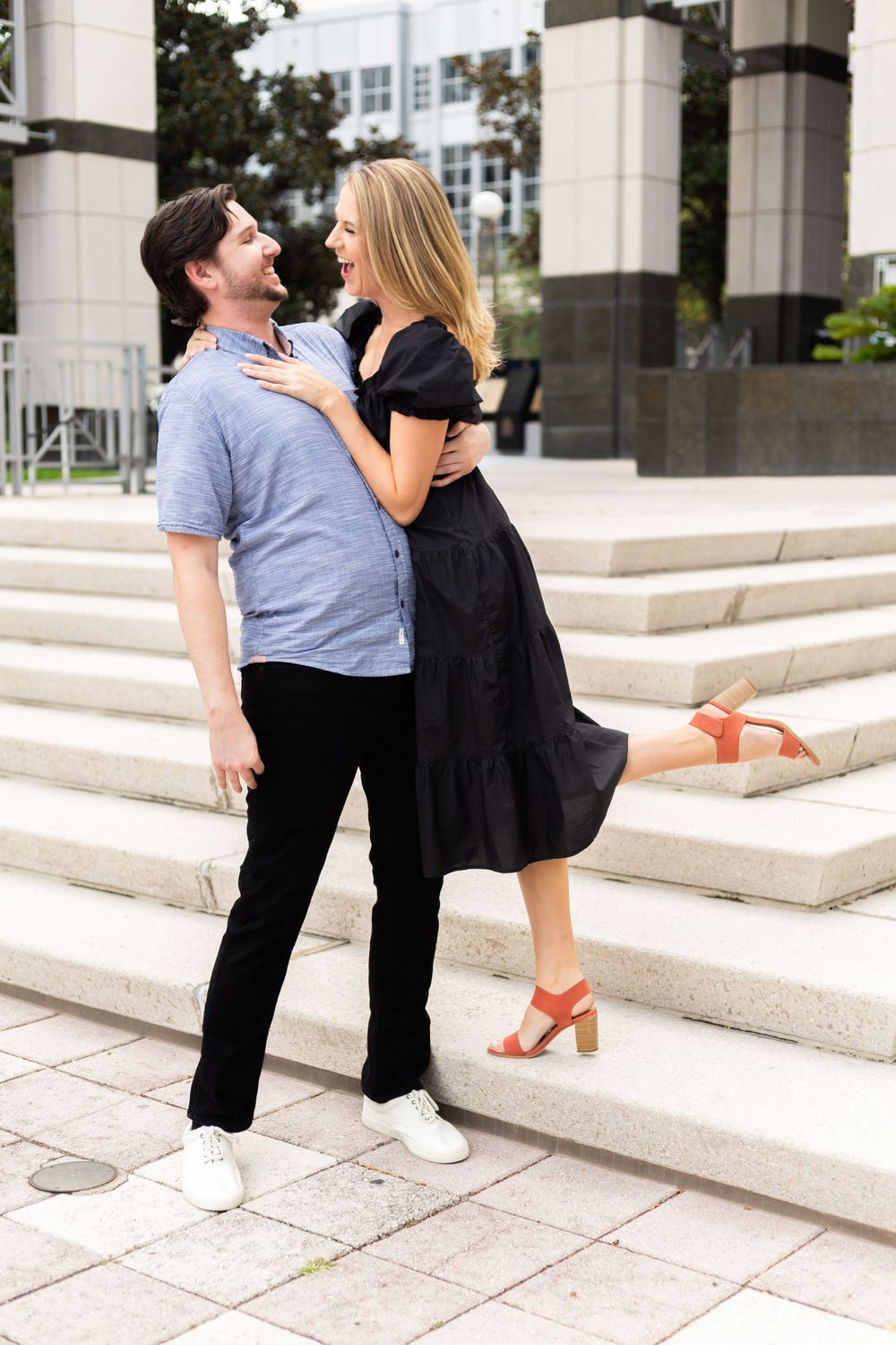 A romantic couple shares a playful moment on outdoor stone steps, with her in a black dress and him in casual blue.