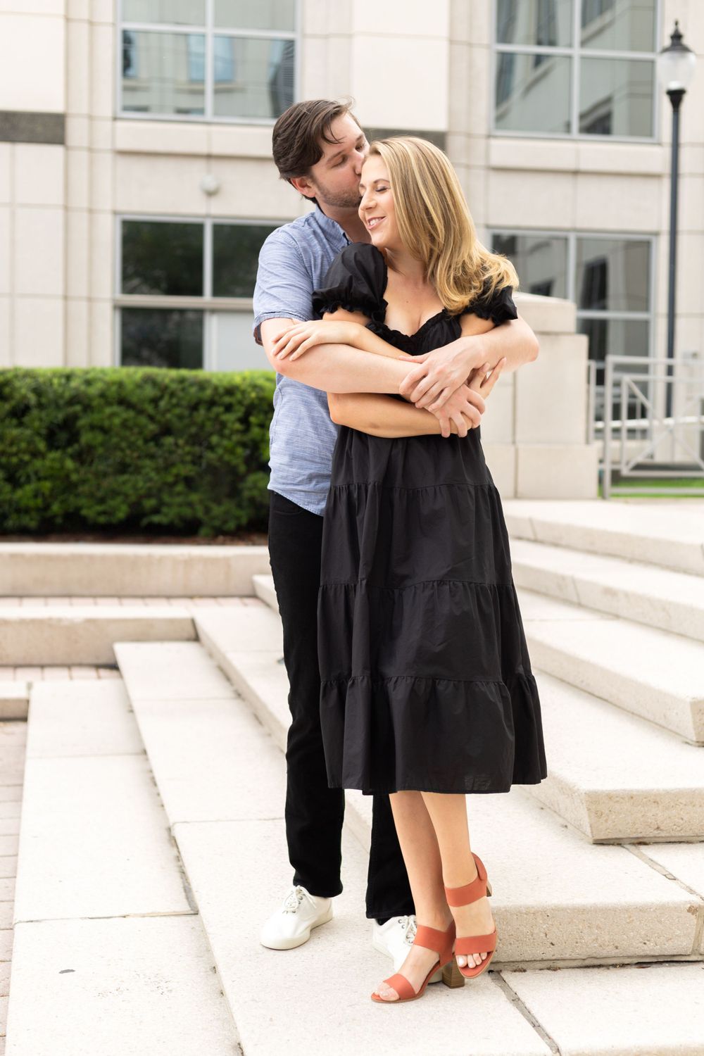 Couple embraces tenderly on white stone steps outside a modern building on a sunny day.