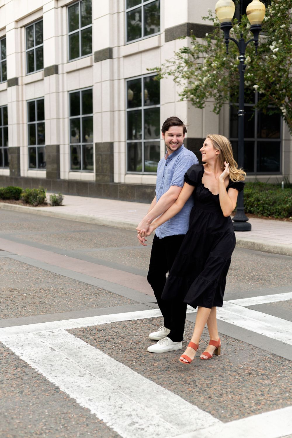 A couple in casual attire dancing together on a crosswalk near a modern building with glass windows.