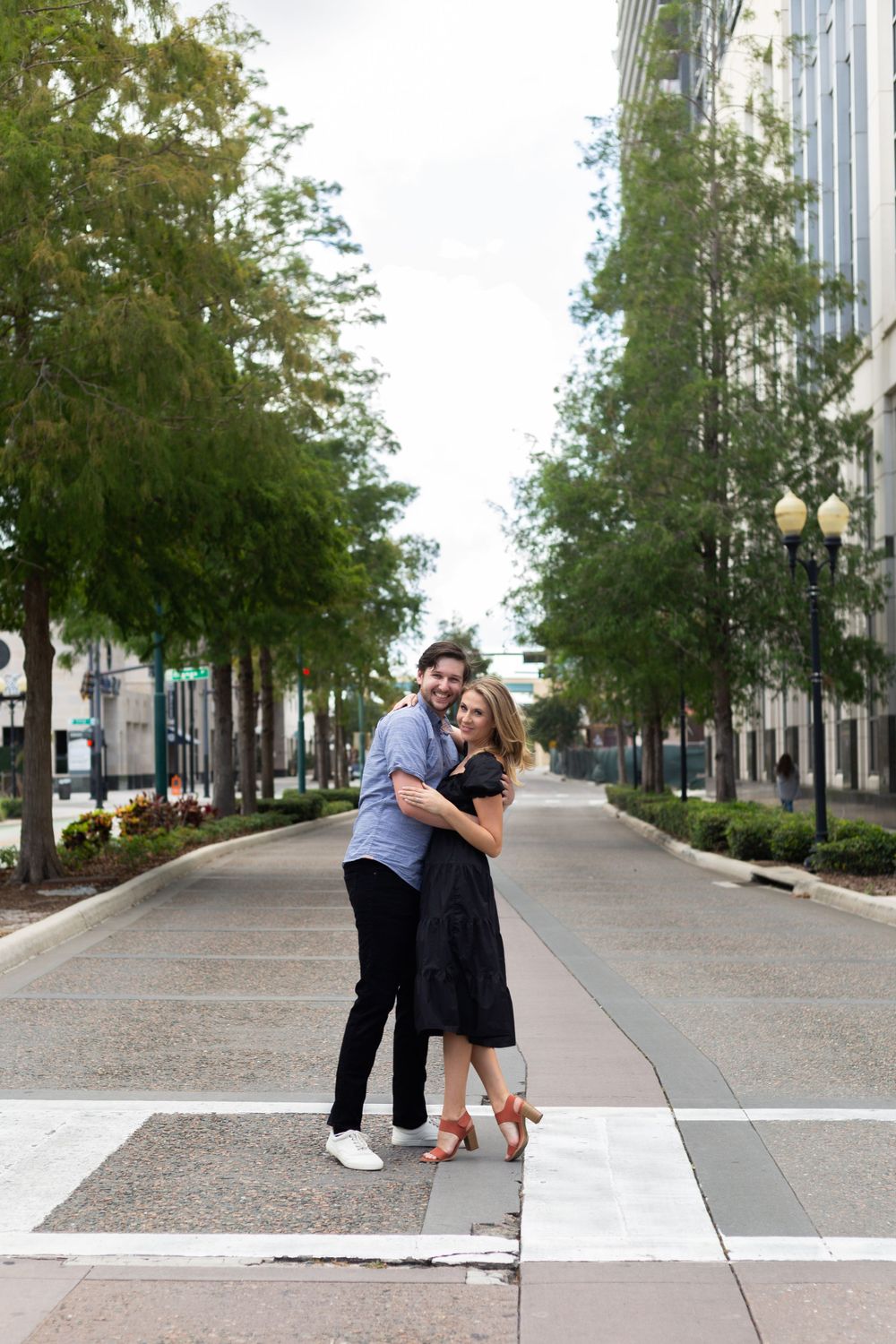 A couple embracing on a tree-lined urban street with decorative lamp posts.