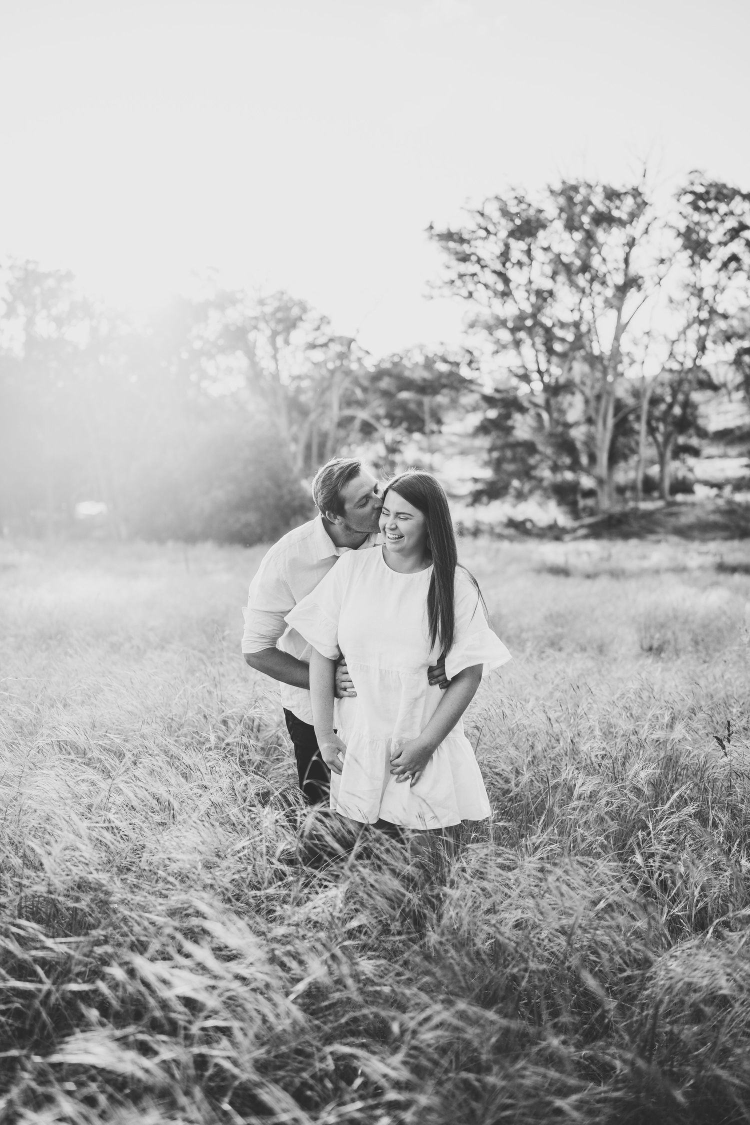 Black and white photo of a couple embracing in a field, man kissing woman on cheek at golden hour.