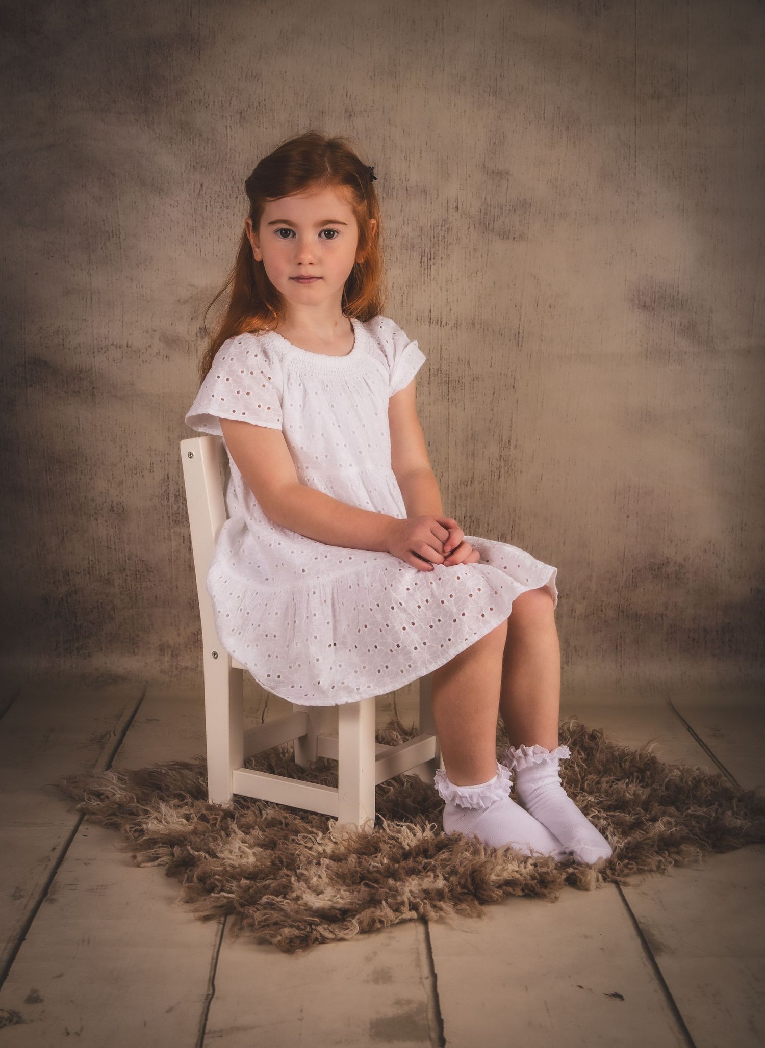 Young child in white eyelet dress poses on chair against textured brown backdrop.