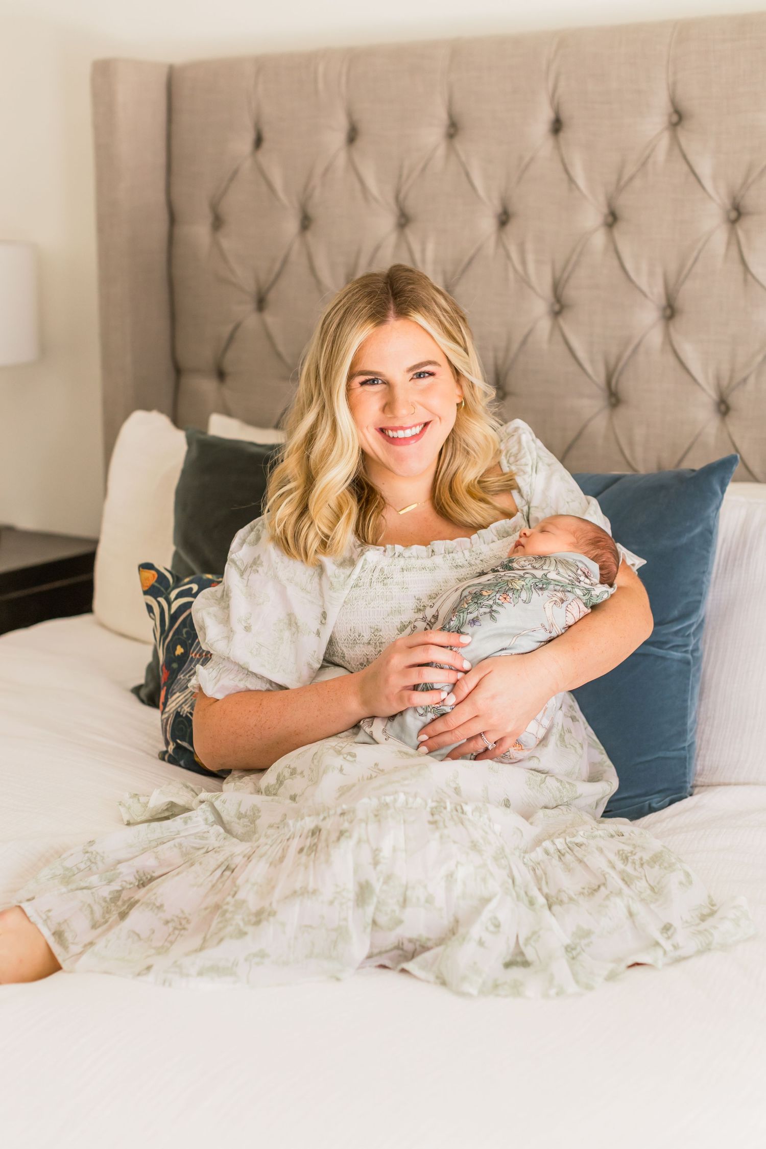 A mother sits in bed holding her newborn baby while wearing a floral dress.