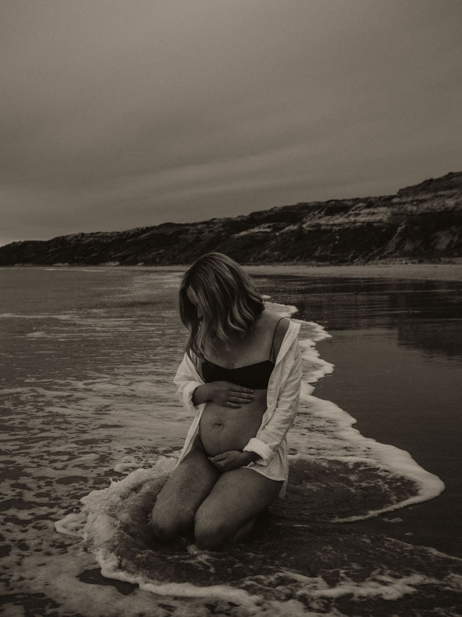 Moody black and white photo of person kneeling in ocean waves near cliffs.