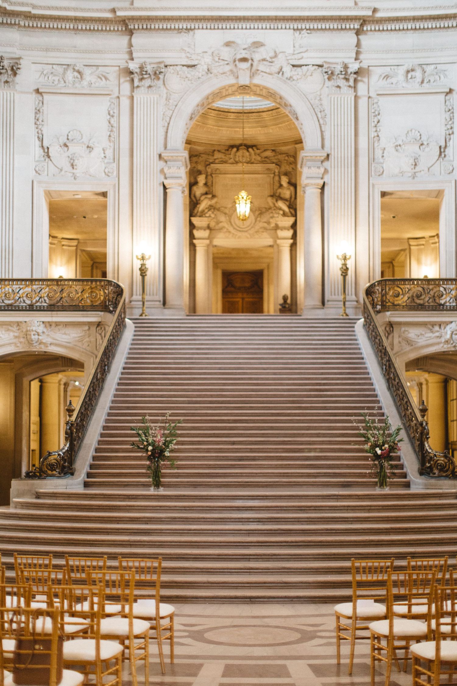 Elegant grand staircase with ornate white marble columns and railings in a classic wedding venue illuminated by chandeliers.