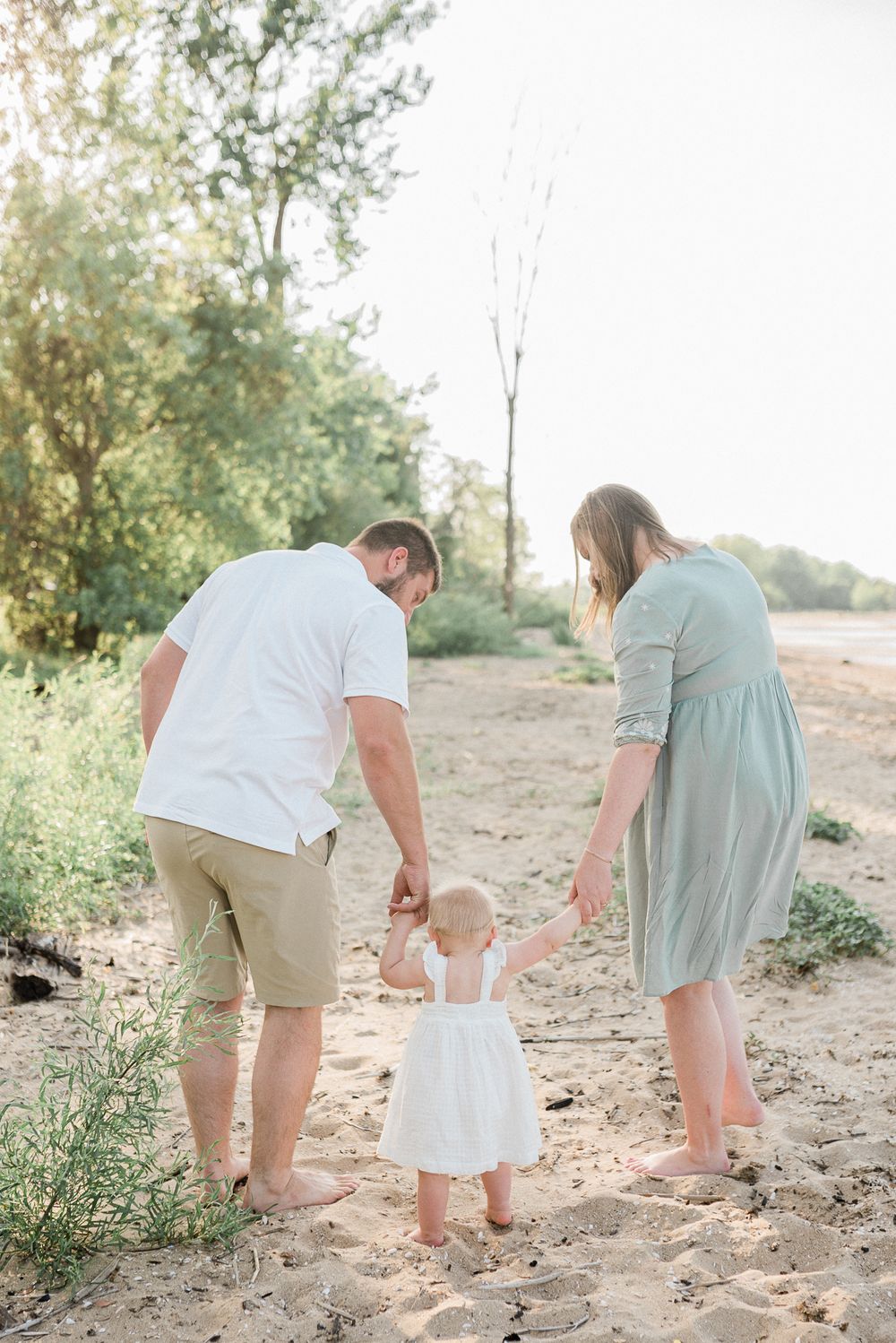 Parents holding hands with their toddler while walking on a sandy path.