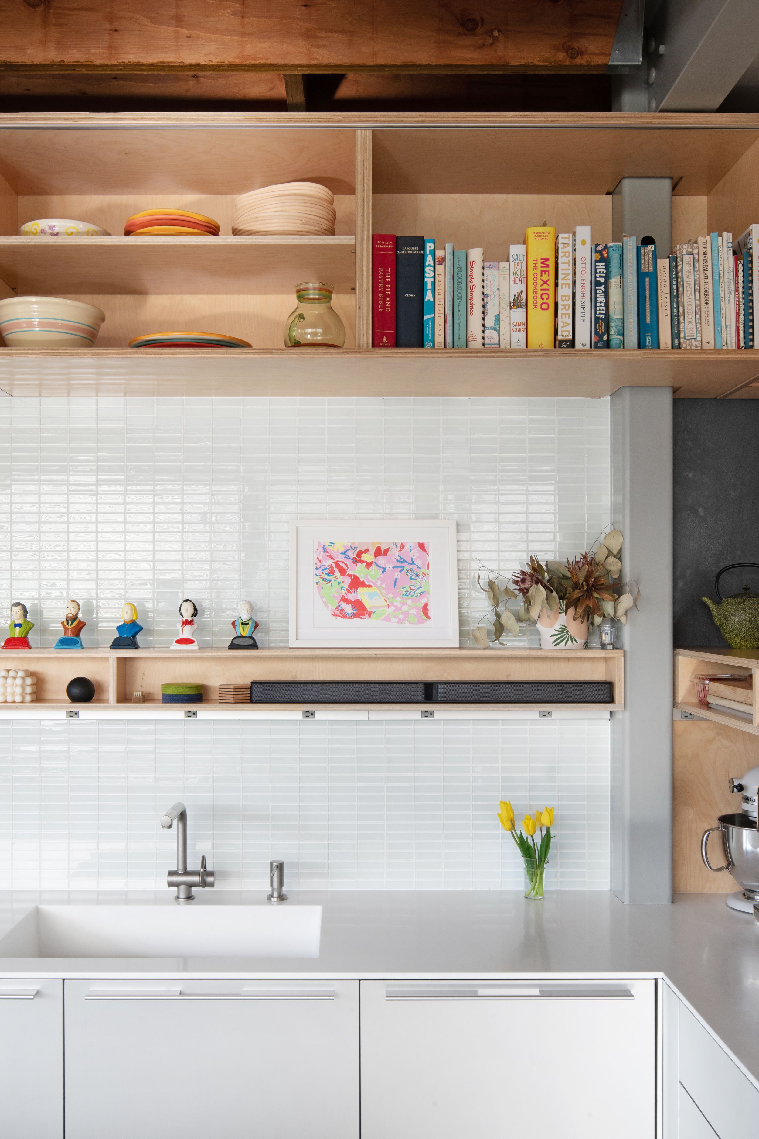 Bright kitchen interior with white cabinets, open shelving displaying books and dishes, and subway tile backsplash.