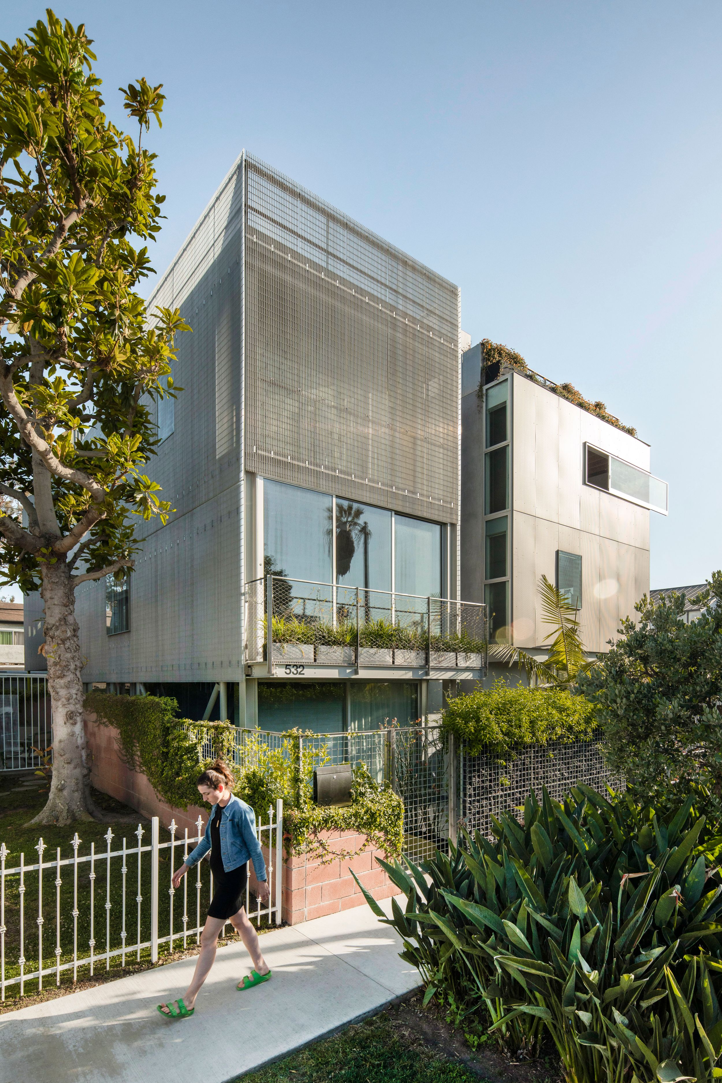 Contemporary multi-level house with glass facade and vertical metal screens surrounded by lush landscaping.