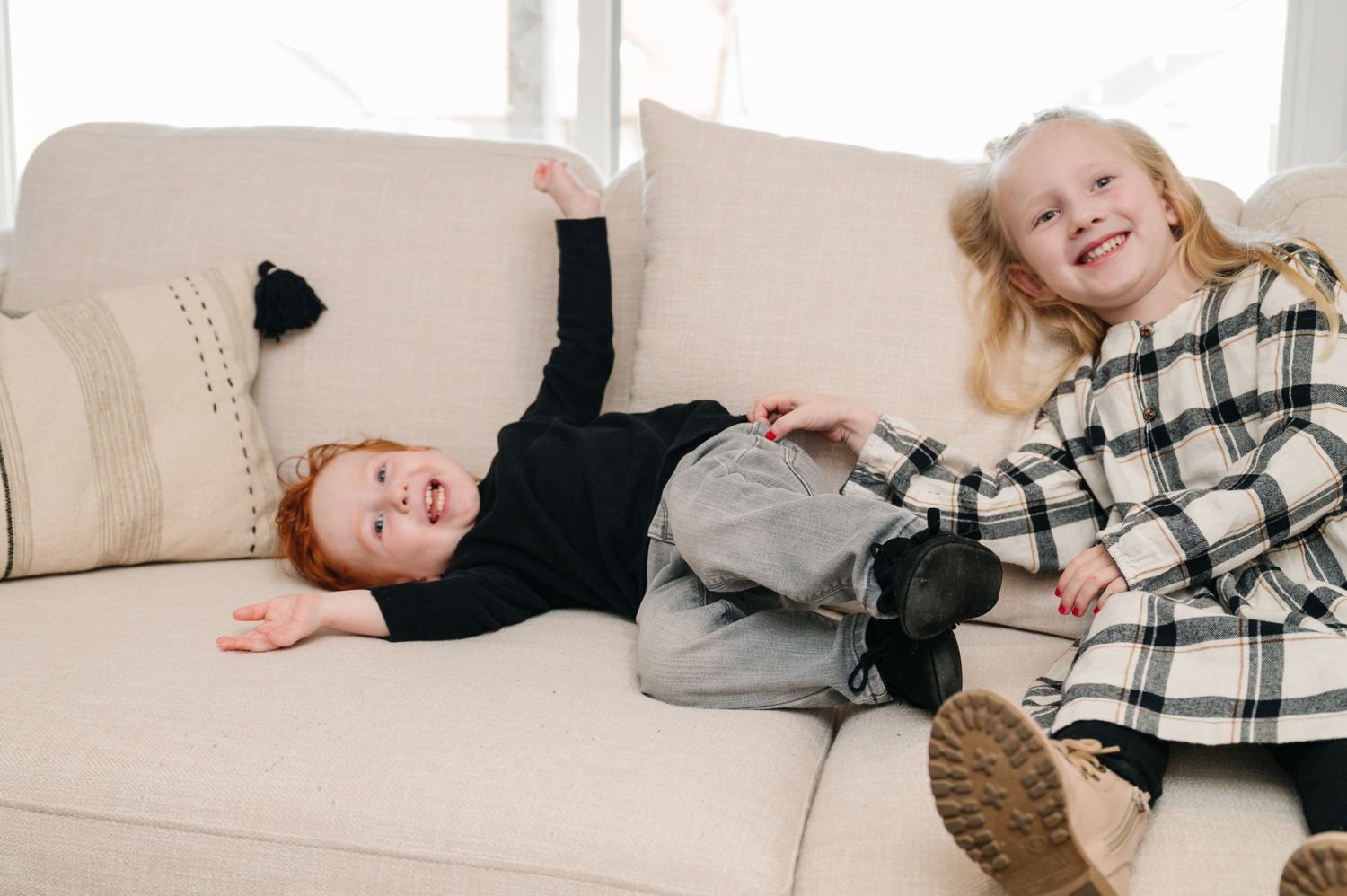Two children playing and laughing on beige couch in casual clothes.