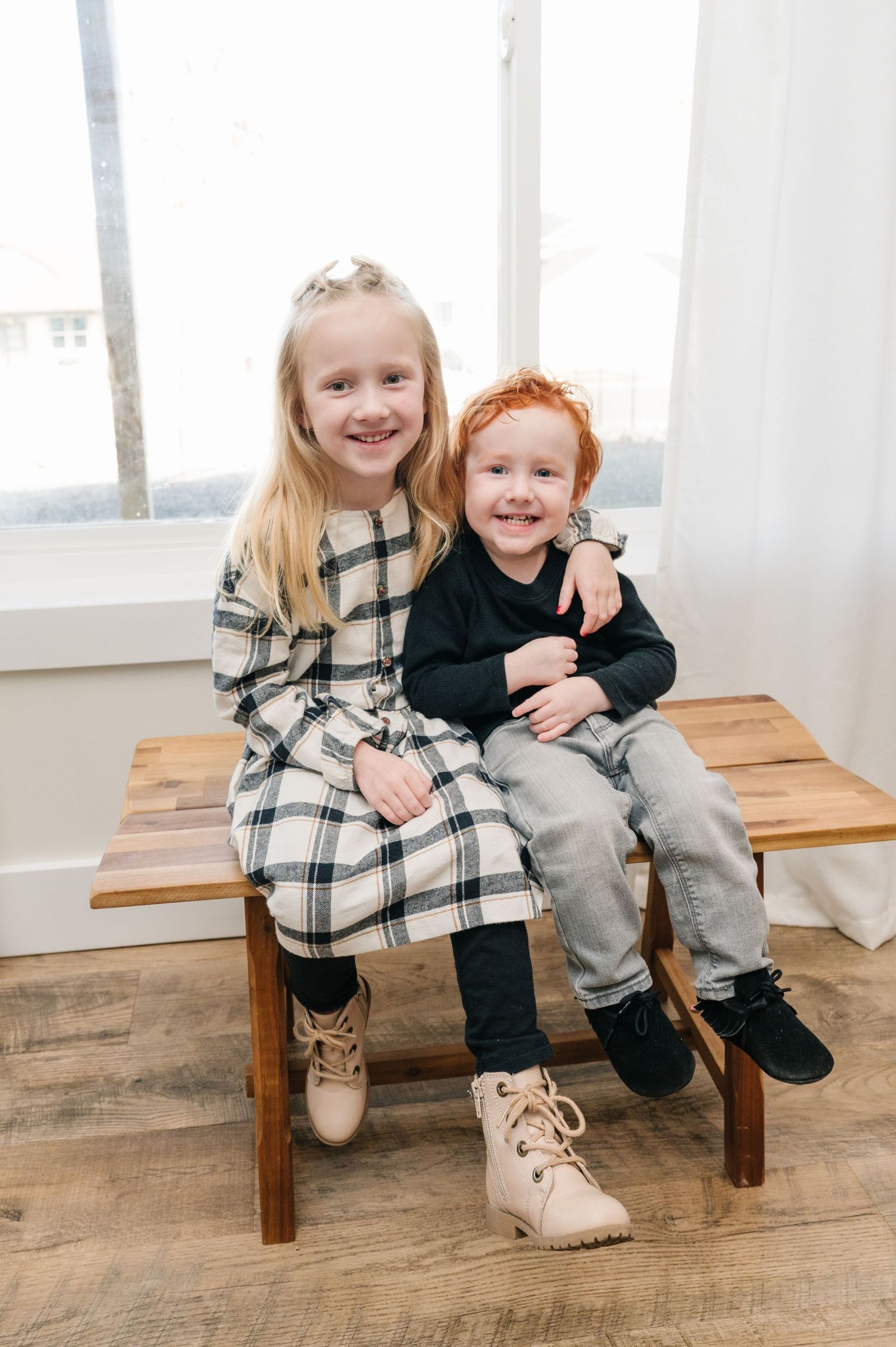 Two young siblings sit together on a wooden bench wearing coordinated winter outfits.