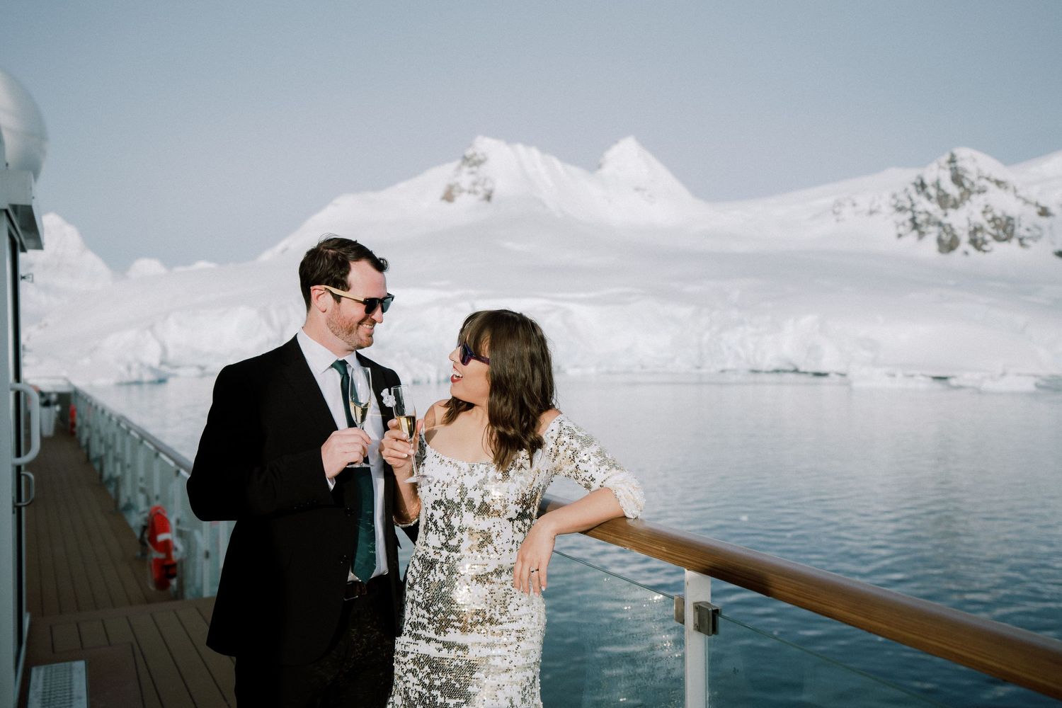 A couple sharing champagne on a cruise ship deck with glaciers and mountains in the background.
