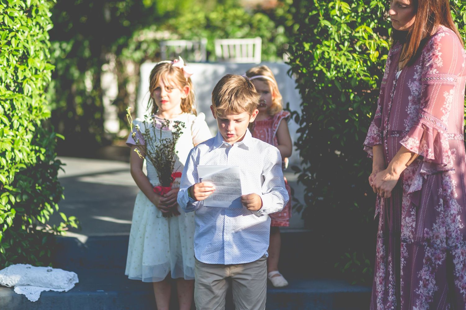 Children standing between green hedges during an outdoor ceremony.