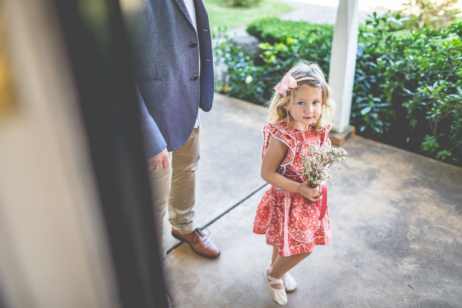 Young girl in floral dress holding flowers on a covered porch.
