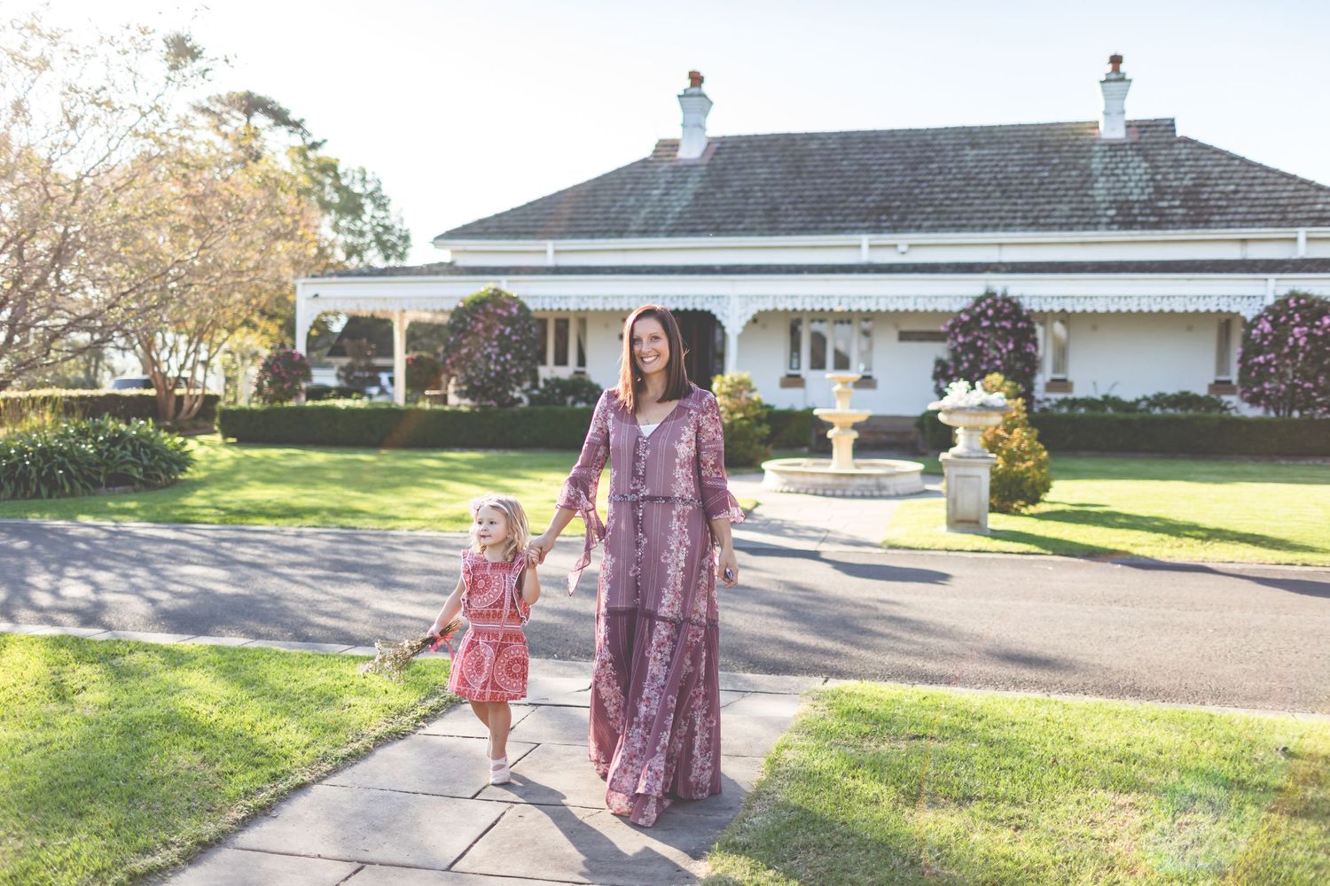 A mother and child walking together on a pathway in front of a white house with manicured lawn.