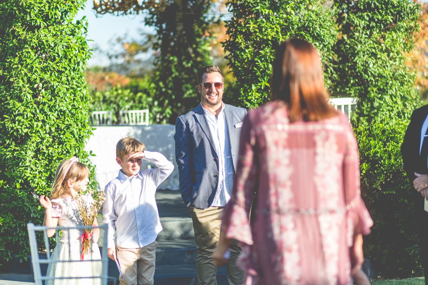 A small outdoor gathering on a sunny day with children and adults meeting near green hedges.