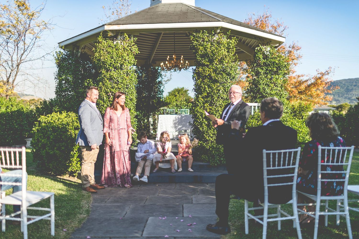 An outdoor ceremony taking place under a vine-covered gazebo with white chairs set up for guests.