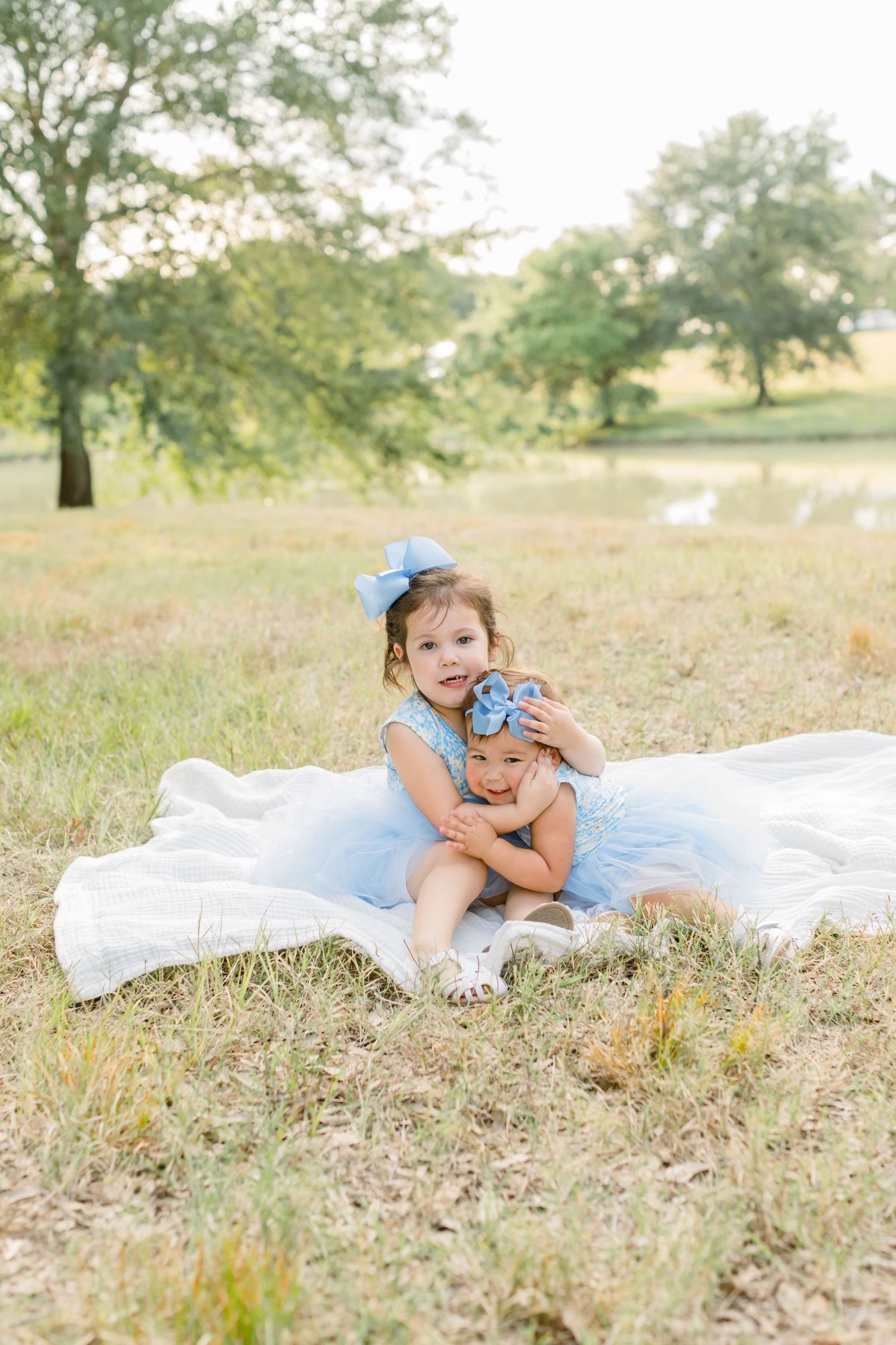Two children in blue dresses sharing sweet moment on white blanket outdoors.