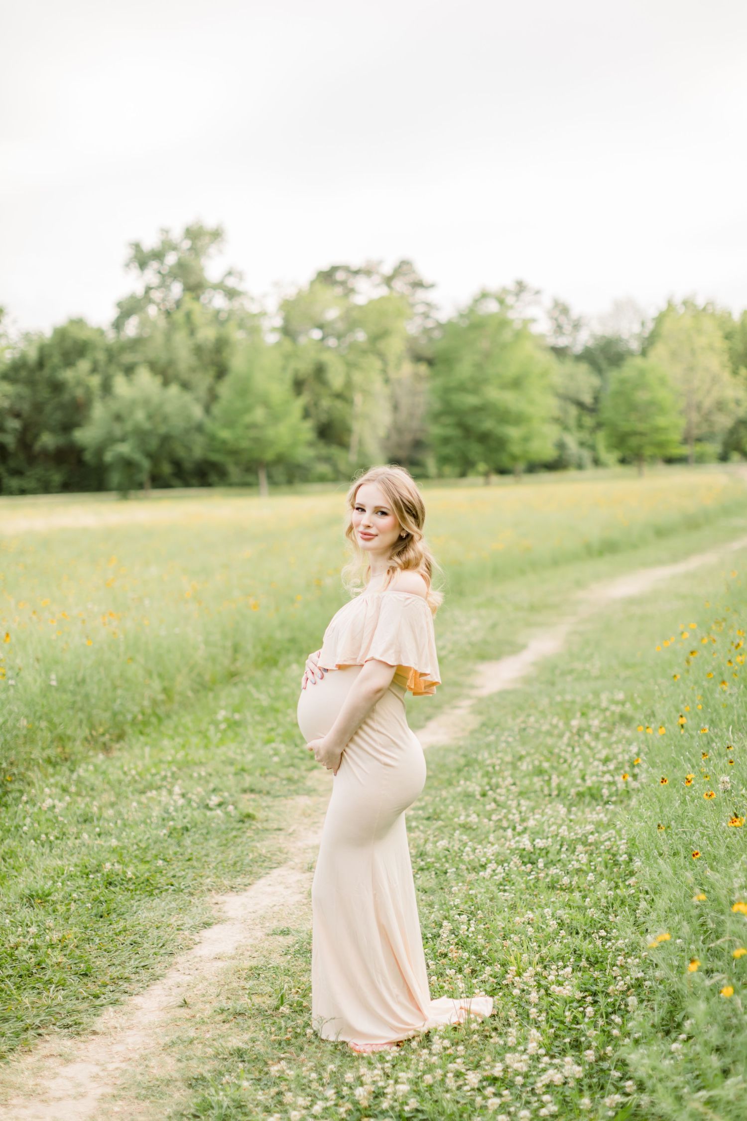 A graceful maternity photo featuring a blush off-shoulder gown in a spring meadow.
