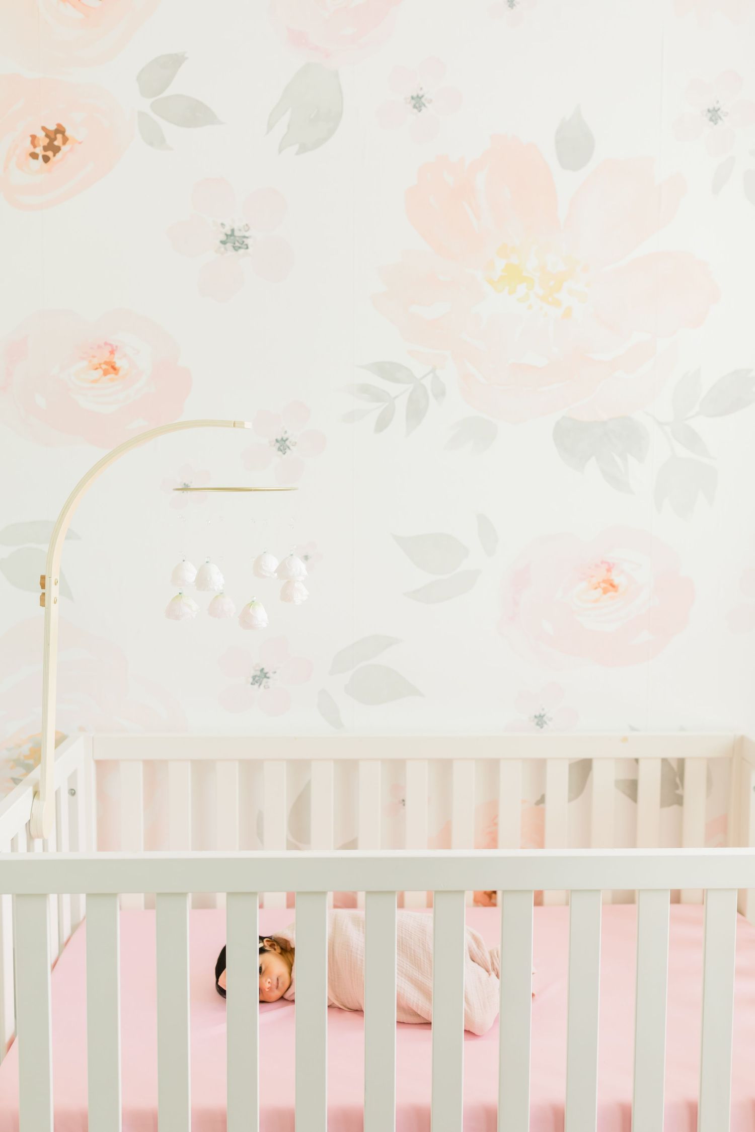 White crib with gold mobile against delicate floral wallpaper in soft pink tones.