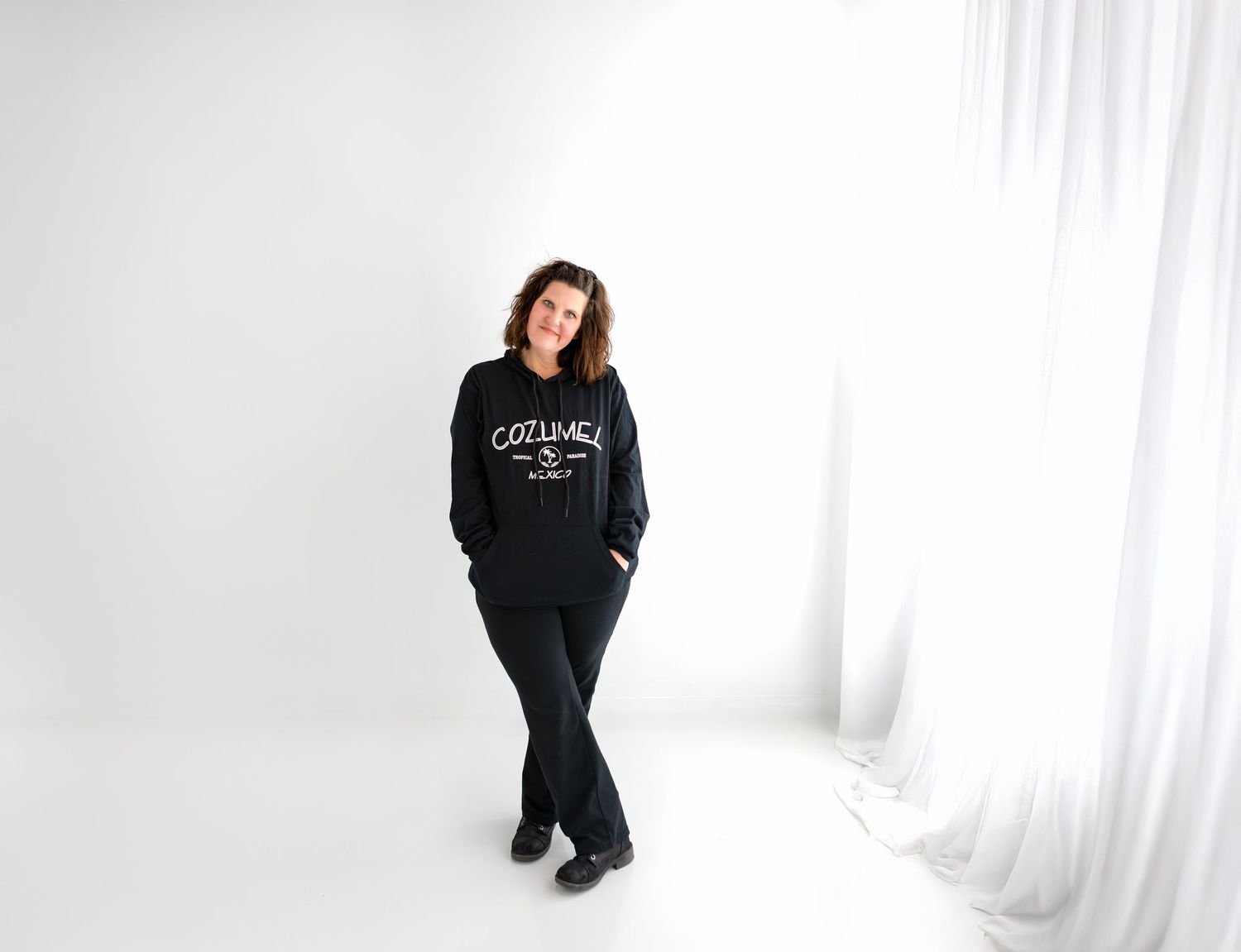 This is Jennifer Cosby the owner of Grit and Grace Photography standing in black clothes in front of a white wall.
