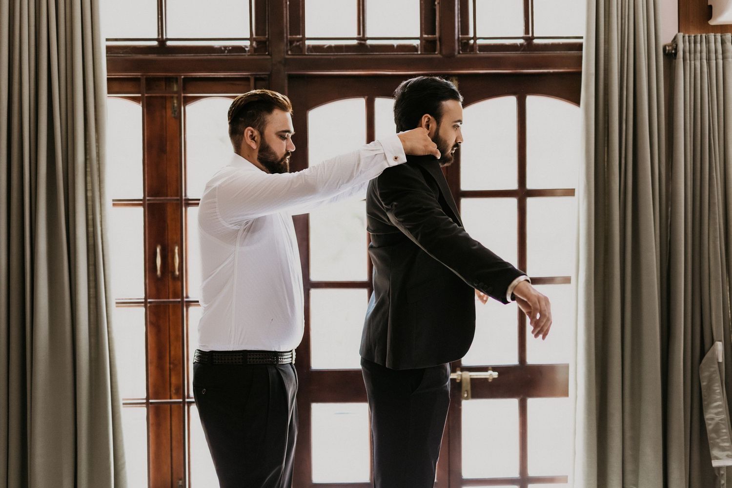 Two people in formal attire stand by large windows during a fitting session.
