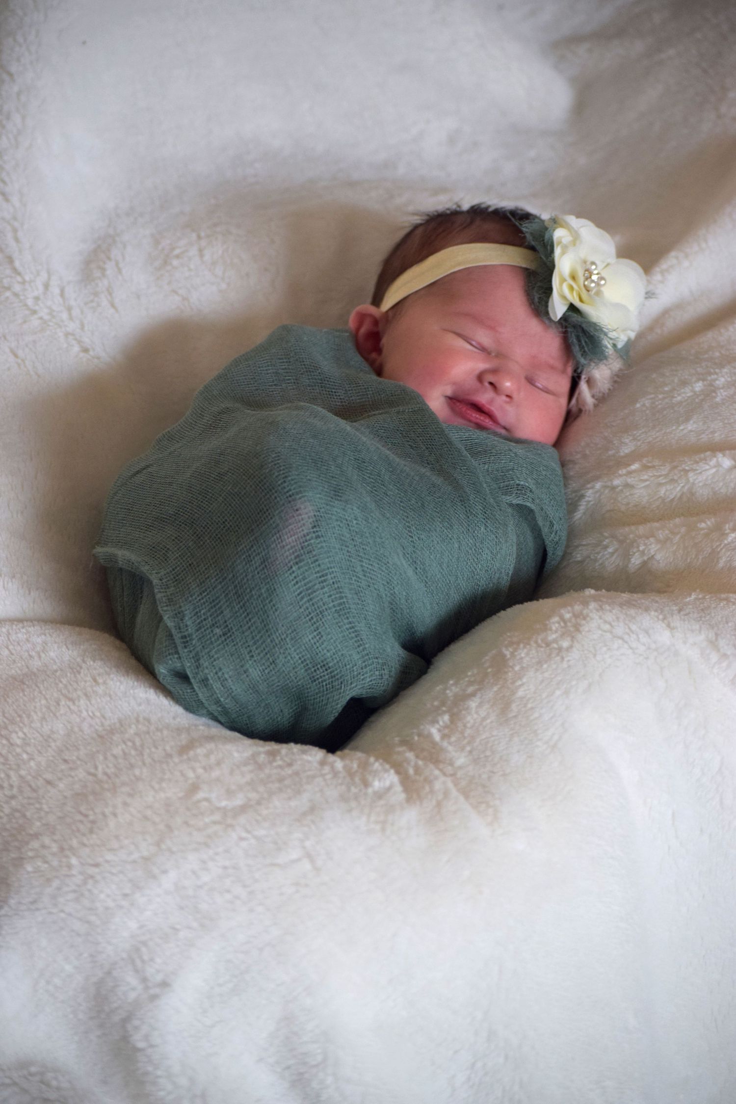 Sleeping newborn wrapped in sage green with delicate floral headband on white blanket.