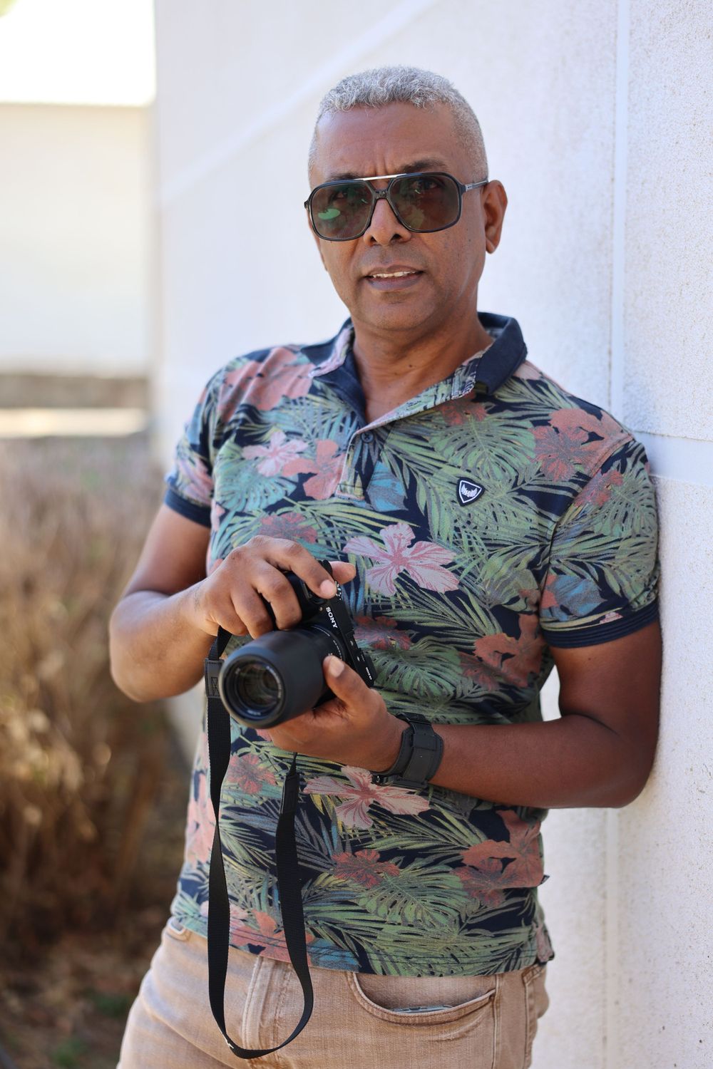 Person wearing a tropical print shirt holding a camera against a white wall.