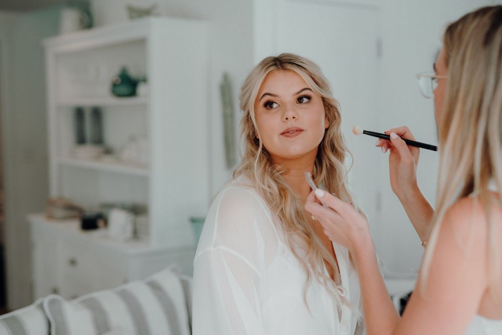 A makeup artist applies cosmetics to a woman in a white robe during wedding preparations.
