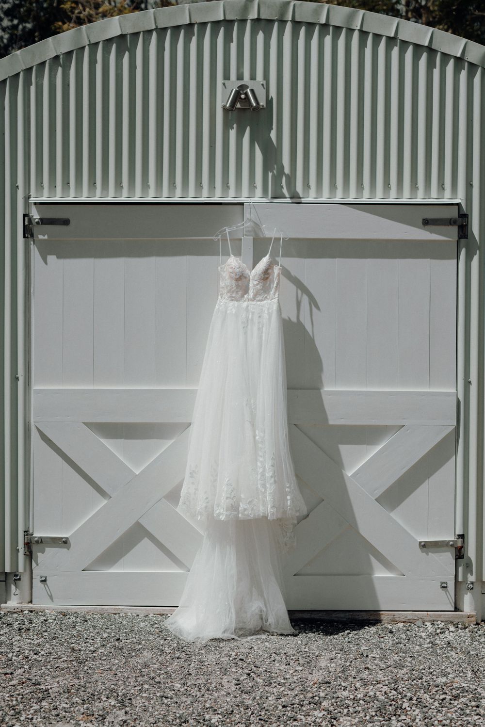 Elegant lace wedding dress hanging on white barn door exterior.