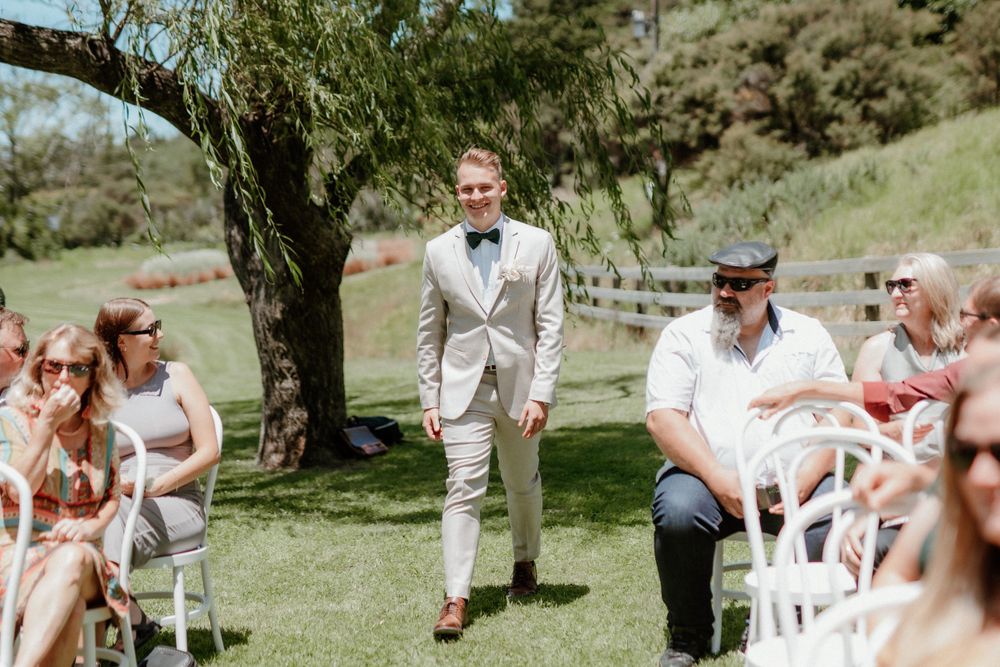 A groom walking down the outdoor ceremony aisle with guests seated on white chairs.