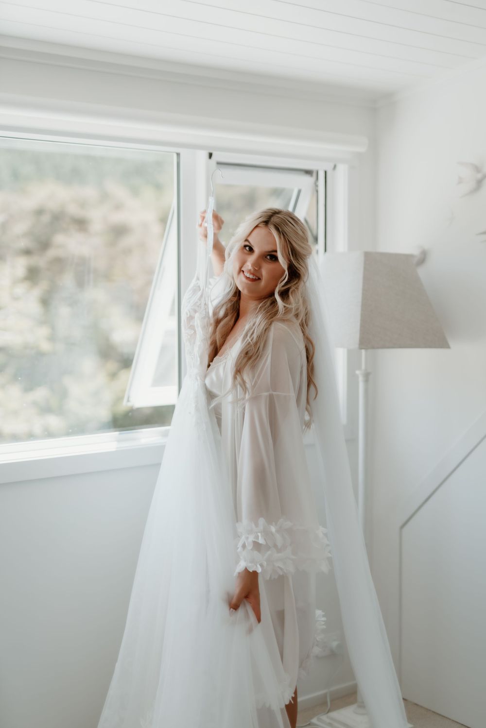 Woman in white robe standing by window with wedding dress.