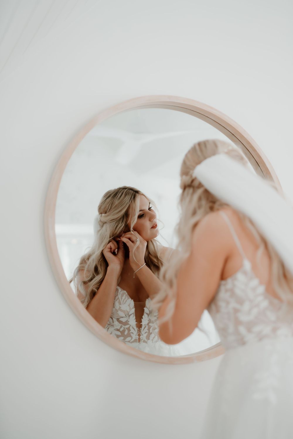 Reflection in round mirror shows bride getting ready in lace dress.